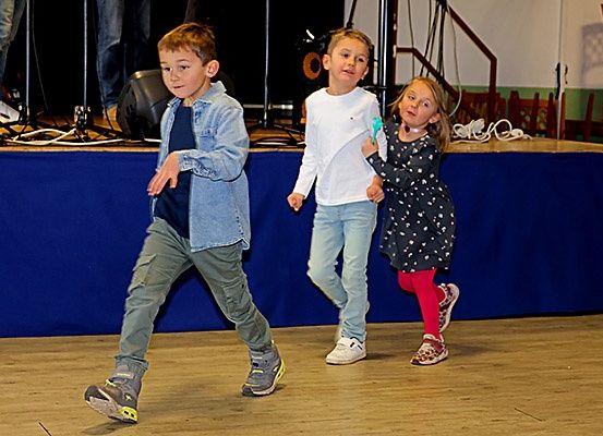 Drei Kinder gehen auf einer Bühne. Der Junge in der Mitte trägt ein weißes T-Shirt. Der Junge auf der linken Seite trägt eine Jeansjacke und Hose. Das Mädchen auf der rechten Seite trägt ein gepunktetes Kleid und Turnschuhe. Hinter ihnen steht ein blauer Tisch auf der Bühne.