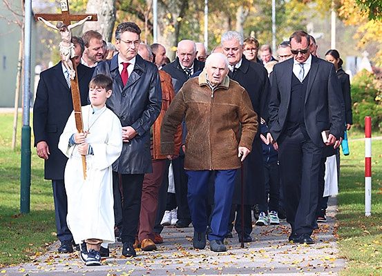Eine Gruppe formell gekleideter Personen geht gemeinsam auf einem Bürgersteig, wobei ein junger Junge ein Kreuz trägt.