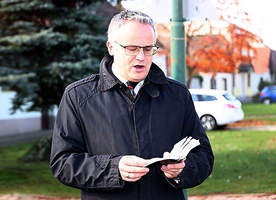 Ein Mann steht draußen in einem Park und liest ein Buch. Er trägt eine dunkle Jacke, eine Krawatte und eine Brille. Hinter ihm befinden sich Bäume, ein Haus und geparkte Autos.