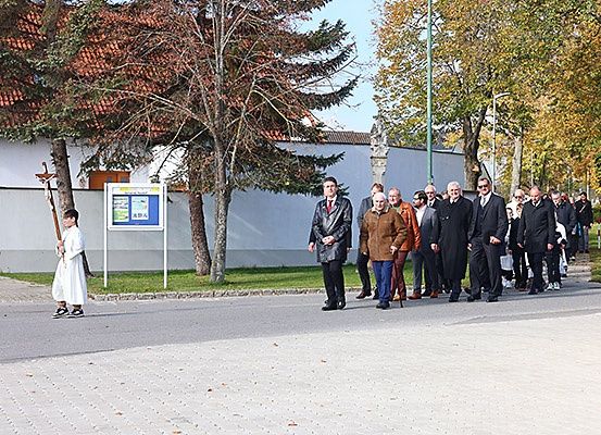 Eine Gruppe von Menschen, formell gekleidet, geht auf einer Straße. Ein junger Mensch in einem weißen Gewand steht links. Ein Schild ist seitlich aufgestellt. Bäume und ein Gebäude sind im Hintergrund.