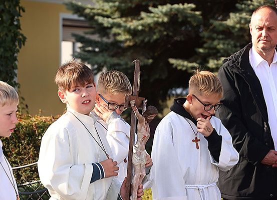 Vier Jungen in weißen Roben halten ein Kruzifix vor einem Gebäude, mit einem Mann in einem schwarzen Mantel und einem weißen Hemd im Hintergrund.