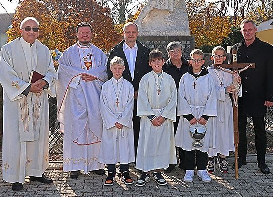 Eine Gruppe von Menschen in weißen Gewändern steht vor einer Statue, darunter Priester, Kinder und ein Mann mit einem Kreuz.