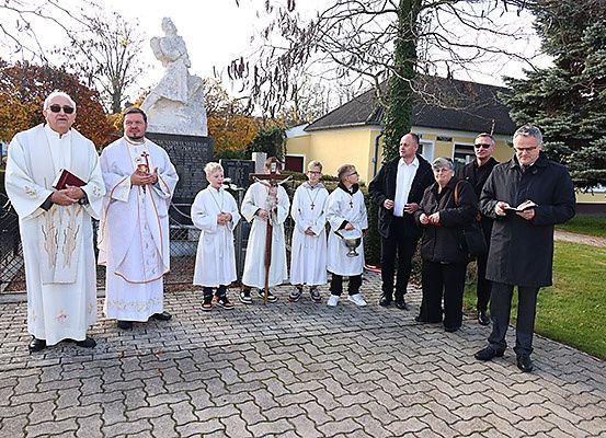 Eine Gruppe von Menschen, darunter Priester und Laien, versammelt sich zu einer Zeremonie vor einer Statue, mit Büchern und einem Kreuz in den Händen.