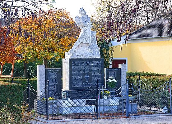 Ein Denkmal in einem Park zeigt eine Statue einer Figur in einem fließenden Gewand, umgeben von einem Zaun mit einem Tor. In der Nähe befindet sich ein gelbes Gebäude mit Grünpflanzen davor.