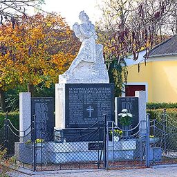 Ein Denkmal in einem Park zeigt eine Statue einer Figur in einem fließenden Gewand, umgeben von einem Zaun mit einem Tor. In der Nähe befindet sich ein gelbes Gebäude mit Grünpflanzen davor.