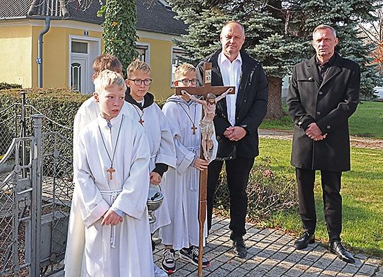 Vier Jungen in weißen Roben, die ein Kreuz halten, stehen mit zwei Männern in Anzügen vor einem Haus.