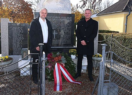 Zwei Männer stehen an einem Denkmal, einer hält einen Kranz, beide lächeln, mit einem Steinmonument und einem Haus im Hintergrund.