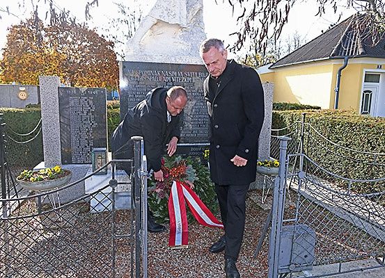 Zwei Männer legen einen Kranz an einem Denkmal mit einer Statue und einem gelben Haus im Hintergrund ab.