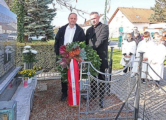 Zwei Männer stehen neben einem Denkmal mit einem Kranz und einer Flagge vor einer Menschenmenge. Ein Gebäude und Autos sind im Hintergrund zu sehen.