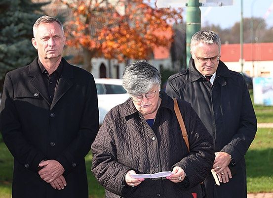 Drei Personen stehen auf einer Rasenfläche, zwei Männer tragen Mäntel und eine Frau hält ein Papier, vielleicht bei einem Trauergottesdienst.