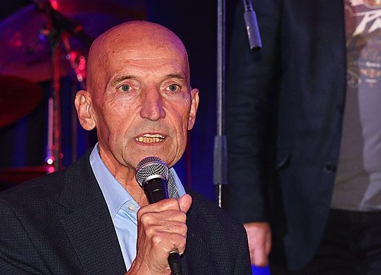 Ein älterer Mann in einem dunklen Anzug spricht in ein Mikrofon auf der Bühne bei einem Konzert.