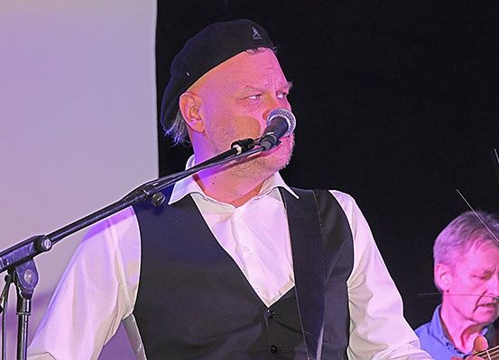 Ein Mann singt in ein Mikrofon auf der Bühne, trägt einen schwarzen Hut und eine Weste. Hinter ihm spielt ein anderer Mann Gitarre.