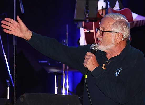 Ein älterer Mann mit Brille und Bart singt auf der Bühne. Er hält ein Mikrofon mit der rechten Hand. Hinter ihm spielt ein Gitarrist.