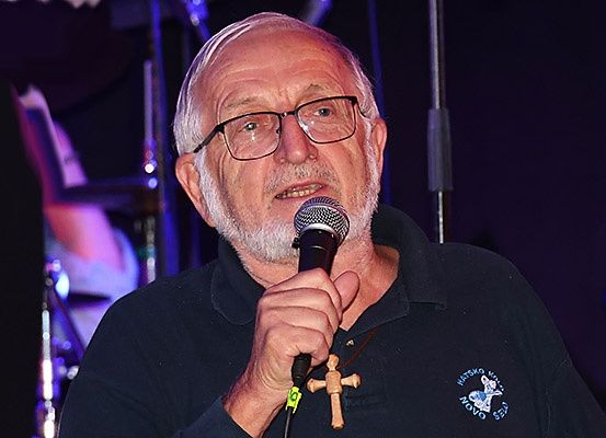 Ein älterer Mann mit Brille und Bart hält ein Mikrofon und spricht in dieses hinein, während er ein Polohemd mit Logo und eine Kreuzkette trägt. Im Hintergrund sind verschwommen ein Schlagzeug und andere Instrumente zu sehen.