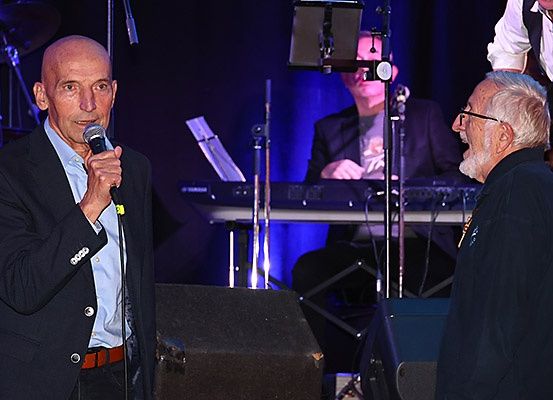 Zwei Männer auf der Bühne, einer mit Mikrofon, der andere mit Brille, und ein Pianist dahinter.