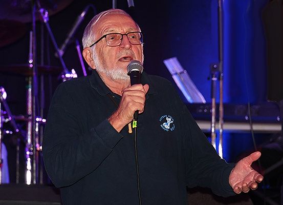 Ein älterer Mann mit Brille spricht in ein Mikrofon auf einer Bühne. Er trägt ein schwarzes Shirt mit einem Logo. Hinter ihm steht ein Schlagzeug und andere Musikinstrumente.