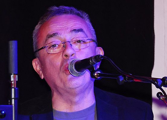 Ein Mann singt in ein Mikrofon unter einem Scheinwerfer, trägt eine Brille und einen dunklen Mantel, mit einem Mikrofonständer und blauem Licht auf der Bühne.