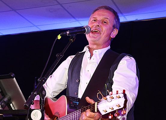 Ein Mann tritt mit einer Gitarre und einem Mikrofon auf der Bühne auf, lächelt und spielt Musik.