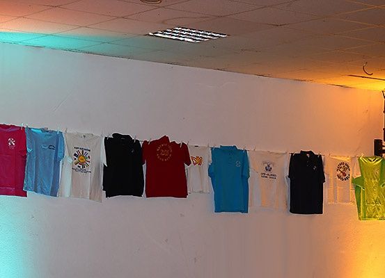 Eine Reihe verschiedener farbiger T-Shirts ist an einer weißen Wand aufgehängt. Jedes Shirt hat ein einzigartiges Design. Die Wand ist weiß gestrichen, und die Decke hat ein quadratisches Fliesendesign. Ein Lautsprecher ist an der Wand montiert.