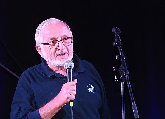 Ein älterer Mann mit Brille hält ein Mikrofon und steht vor einem Mikrofonständer, mit einem Logo auf seinem Shirt.