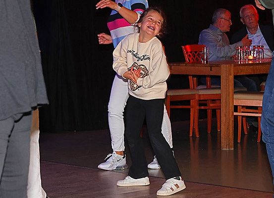 Ein junges Mädchen tanzt mit einer Frau vor einem Tisch, an dem zwei Männer sitzen.