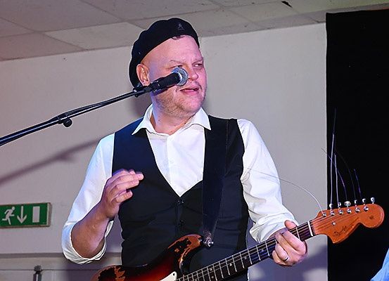 Ein Mann in schwarzer Mütze und Weste singt in ein Mikrofon und spielt Gitarre auf der Bühne.