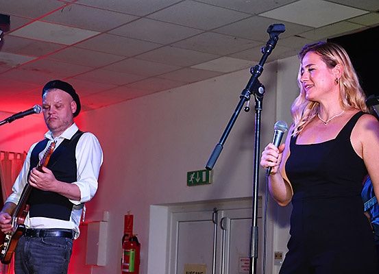 Ein Mann spielt Gitarre und eine Frau singt in ein Mikrofon auf einer Bühne mit roter Beleuchtung.