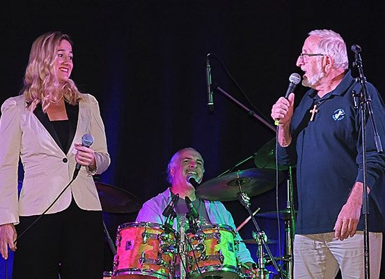 Drei Personen sind auf der Bühne und treten auf. Ein Mann und eine Frau singen, während ein anderer Mann Schlagzeug spielt.
