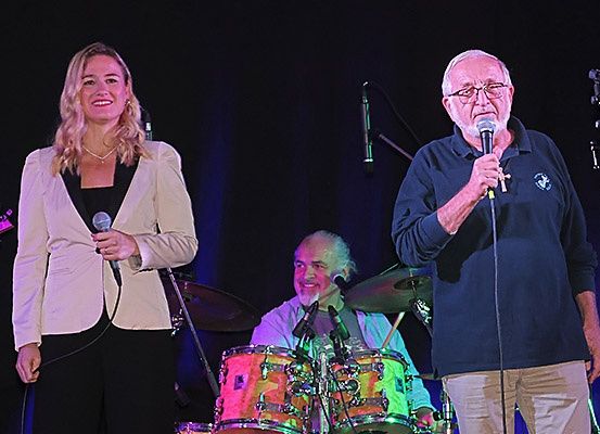 Eine Frau und ein Mann stehen mit einem Schlagzeuger auf der Bühne, alle halten Mikrofone und treten unter Bühnenlicht auf.