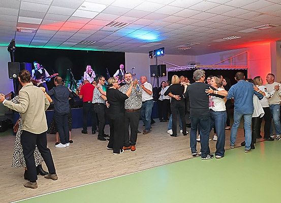 Eine Band spielt auf der Bühne mit farbigen Lichtern, während Menschen in einem Saal mit Holzböden und grünem Boden tanzen.