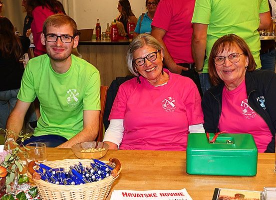 Drei Personen sitzen an einem Tisch mit einer grünen Box und einem Korb mit Stiften und lächeln. Die Frau in der Mitte trägt ein rosa Shirt, während der Mann auf der linken Seite ein grünes Shirt trägt. Der Tisch hat eine Pflanze, ein Glas und ein Buch. Im Hintergrund stehen Personen, einige tragen Brillen.