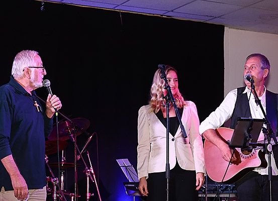 Drei Musiker treten auf der Bühne auf, ein Mann links hält ein Mikrofon, eine Frau in der Mitte singt und ein Mann rechts spielt Gitarre. Hinter ihnen steht ein Schlagzeug und ein Notenständer.