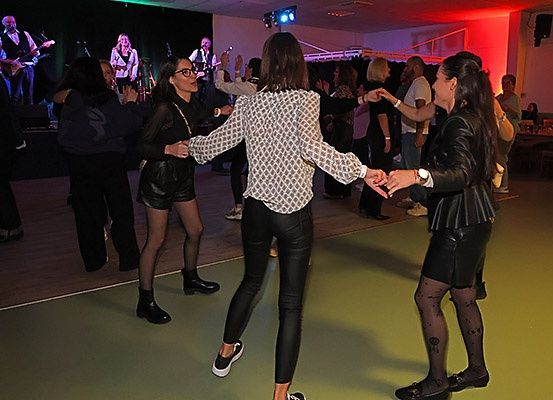 Eine Gruppe von Menschen tanzt zusammen auf einer Bühne, während eine Band im Hintergrund spielt. Die Tänzer umfassen zwei Frauen, die sich an den Händen halten, und eine Frau mit Brille.