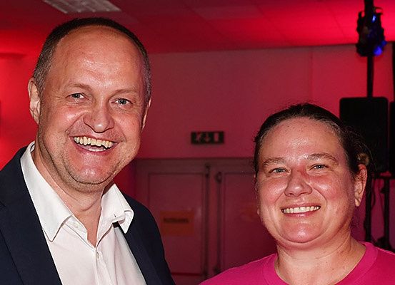 Ein Mann und eine Frau lächeln und posieren für ein Foto. Sie tragen beide formelle Kleidung. Hinter ihnen befindet sich eine rote Wand mit einem Schild und einer Tür. Es gibt auch Lautsprecher und eine Lampe an der Decke.