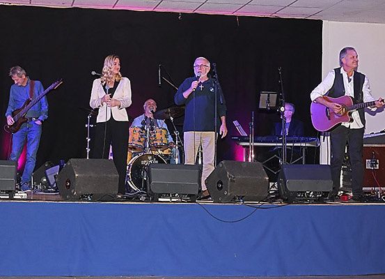 Eine Band spielt auf der Bühne mit einem Schlagzeuger, einem Gitarristen und einem Keyboarder. Ein Mann mit einem Mikrofon singt vor einer Frau.