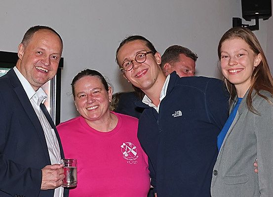 Vier Personen, zwei Männer und zwei Frauen, lächeln und posieren für ein Foto. Der Mann links hält ein Glas, und der Mann neben ihm trägt eine Brille. Die Frau in der Mitte trägt ein pinkes Shirt mit Logo, und die Frau rechts trägt einen grauen Blazer.