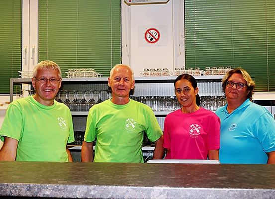 Vier Personen in passenden Shirts stehen hinter einer Bar. Drei sind in Grün und eine in Pink. Die Bar ist vor einem Hintergrund aus grünen Jalousien und einem Rauchverbotsschild platziert.
