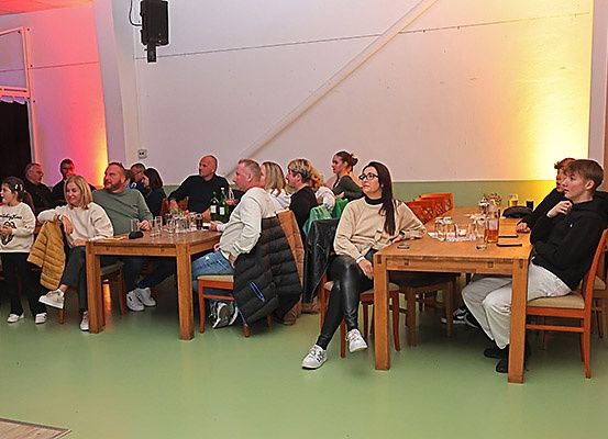 Eine Gruppe von Menschen sitzt an Tischen in einem Raum mit grünem Boden und weißen Wänden, einige mit Brille und Turnschuhen. Tische haben Gläser und Flaschen. An der Wand ist ein Lautsprecher montiert.