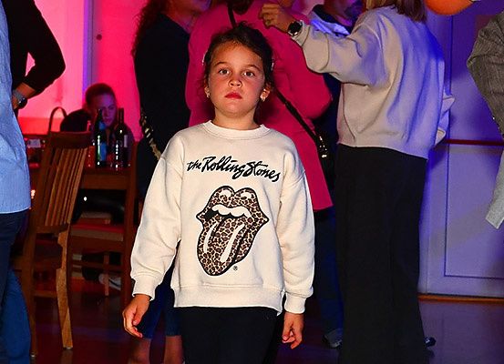 Ein junges Mädchen in einem Rolling Stones Sweatshirt steht in einem Raum mit rosa Beleuchtung, umgeben von Menschen.