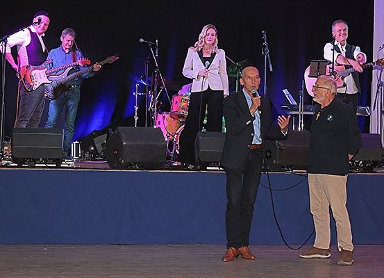 Eine Band tritt auf einer Bühne mit blauem Hintergrund auf. Zwei Männer und eine Frau spielen Gitarren, und ein Mann in Anzug spricht in ein Mikrofon. Die Bühne ist mit Lautsprechern und Mikrofonen ausgestattet.