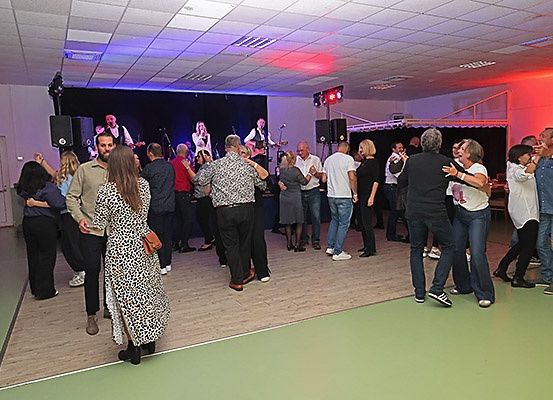 Eine Gruppe von Menschen tanzt in einem Raum mit Bühne, Lautsprechern und bunten Lichtern, einige mit Gitarren und Mikrofonen.