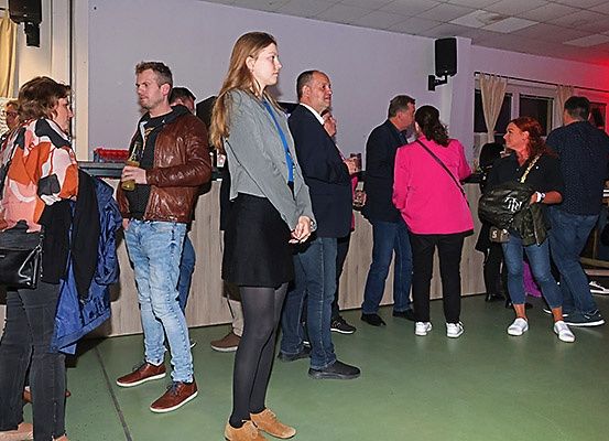 Eine Gruppe von Menschen steht in einem Raum, einige halten Getränke in der Hand, lässig gekleidet. Eine Frau in Schwarz unterhält sich mit einem Mann im braunen Ledermantel.