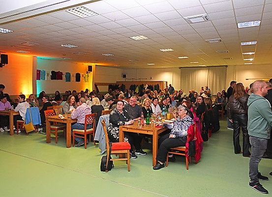 Eine Gruppe von Menschen speist in einem Raum mit grünem Boden. Einige sitzen an Tischen, während andere stehen und sich unterhalten. Kleidung hängt an der Wand und Lautsprecher sind an der Wand montiert.