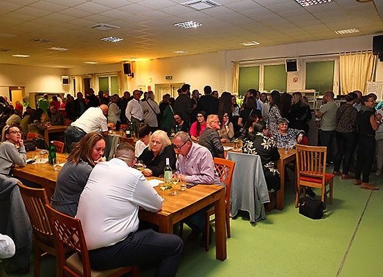Eine große Gruppe von Menschen sitzt an Tischen in einem Saal. Sie scheinen eine Mahlzeit einzunehmen. Die Tische sind mit verschiedenen Gegenständen gefüllt, darunter Gläser, Flaschen und andere Gegenstände. Der Raum hat weiße Wände und grünen Boden. Es gibt Fenster mit Vorhängen und ein Schild ist an der Wand montiert.