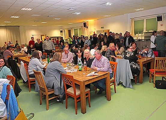 Eine Gruppe von Menschen sitzt um Holztische in einem Essbereich, einige mit Gläsern und Flaschen, während andere in der Nähe der Theke stehen. Der Raum hat weiße Wände, grünen Boden und einen Lautsprecher an der Wand.