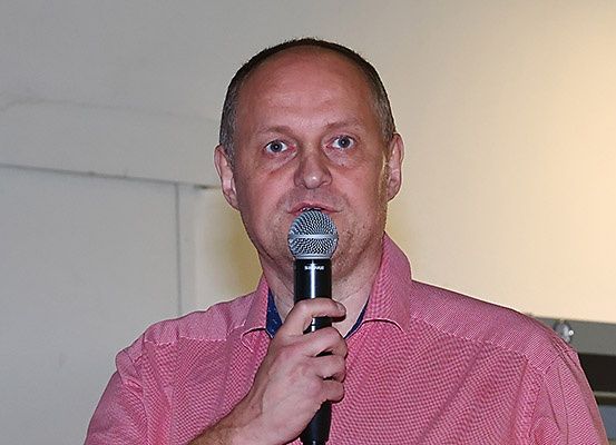 Ein Mann mit kurzen Haaren, der ein pinkes kariertes Hemd trägt, hält ein Mikrofon und spricht in dieses. Der Ort scheint drinnen zu sein, möglicherweise bei einer öffentlichen Veranstaltung.