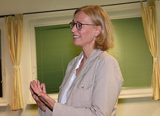 Eine Frau mit kurzen Haaren, die eine Brille und einen hellen Blazer trägt, steht vor einem Fenster, lächelt und klatscht in die Hände.