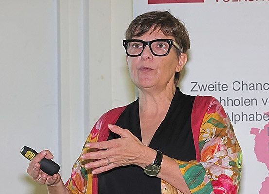 Eine Frau mit Brille und einer Uhr spricht in ein Mikrofon vor einem weißen Banner.