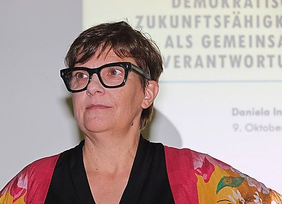 Eine Frau mit Brille steht vor einer Projektionsleinwand mit Text in Deutsch. Sie trägt ein rotes und gelbes Blumentop mit einem schwarzen Innenteil.