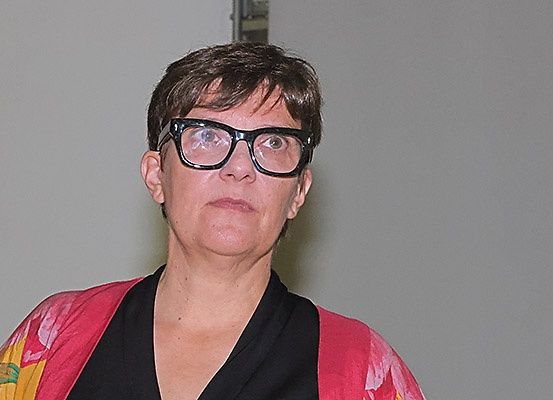 Eine Frau mit kurzen Haaren und Brille steht vor einer weißen Wand und trägt ein rosa und schwarzes Oberteil.